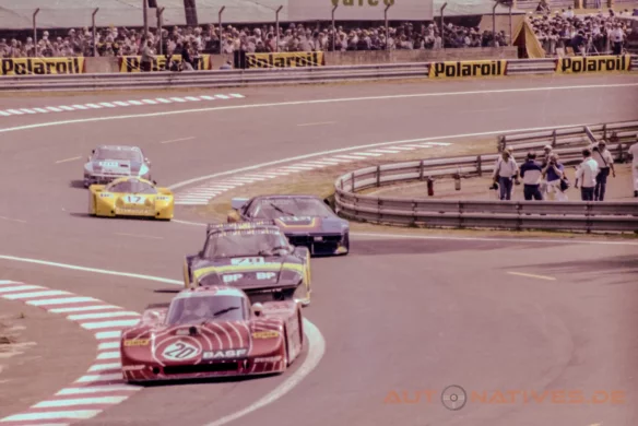 Sauber SHS C6 im rot‑weißen BASF‑Design führt 1982 in der Dunlop‑Schikane von Le Mans eine Gruppe aus Porsche 935 K3, BMW M1, Porsche 924 Carrera GTR und gelber Lola T610 an; Zuschauer und Fotografen säumen die Strecke.