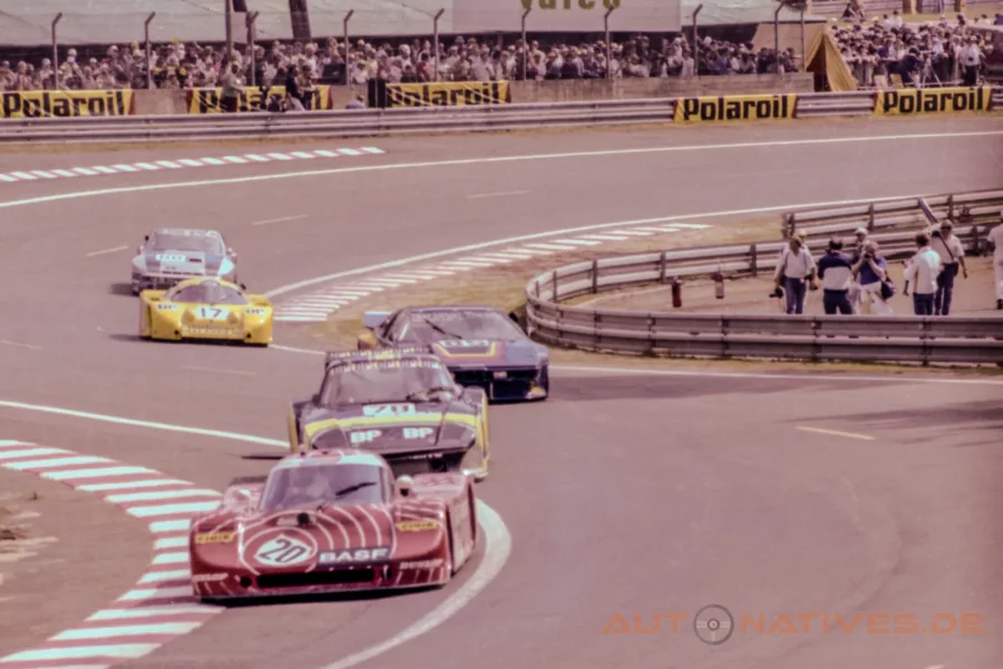 Sauber SHS C6 im rot‑weißen BASF‑Design führt 1982 in der Dunlop‑Schikane von Le Mans eine Gruppe aus Porsche 935 K3, BMW M1, Porsche 924 Carrera GTR und gelber Lola T610 an; Zuschauer und Fotografen säumen die Strecke.